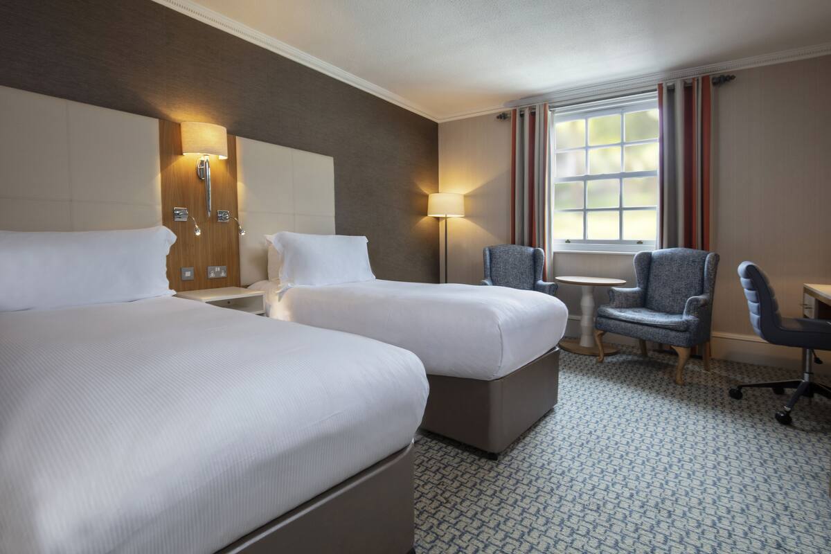 A hotel room with two beds, a desk, a chair, a lamp, and a window with curtains.