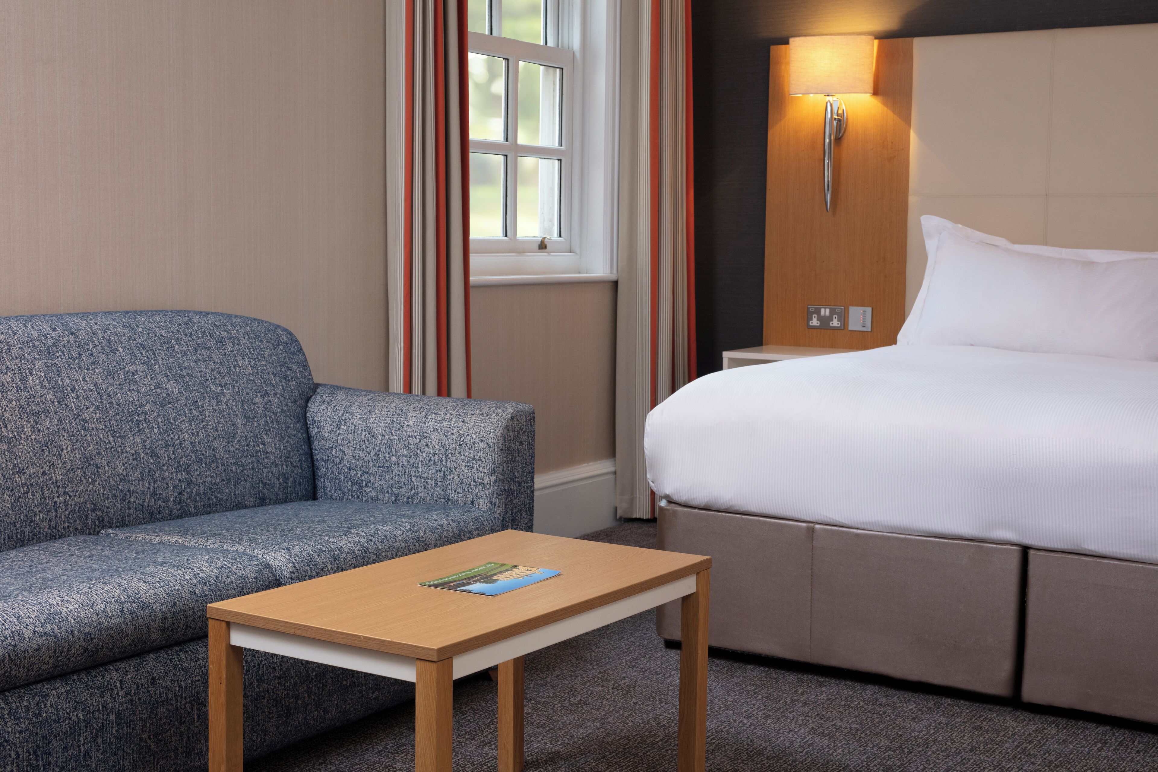 Een hotelkamer met een blauwe bank, een houten tafel, een bed met wit beddengoed en een raam met gordijnen.