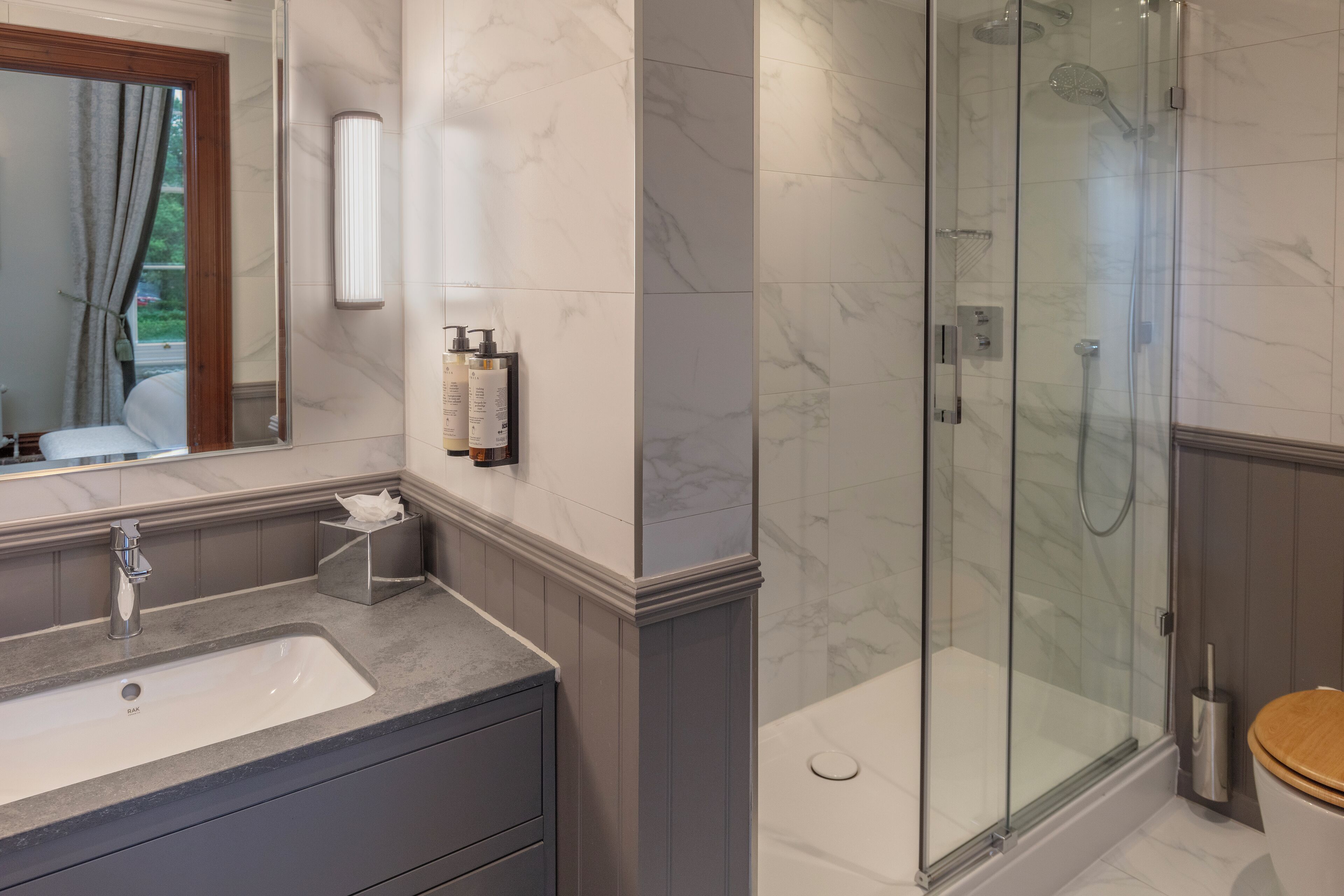 A bathroom with a marble-tiled wall, a glass-enclosed shower, a wooden toilet, and a vanity with a sink and mirror.