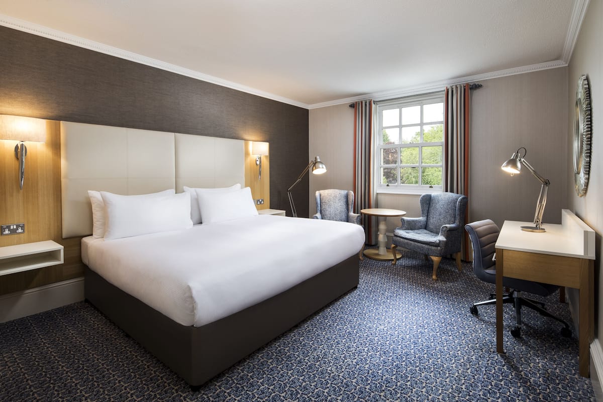 A hotel room with a large bed, two armchairs, a small round table, and a desk with a lamp.
