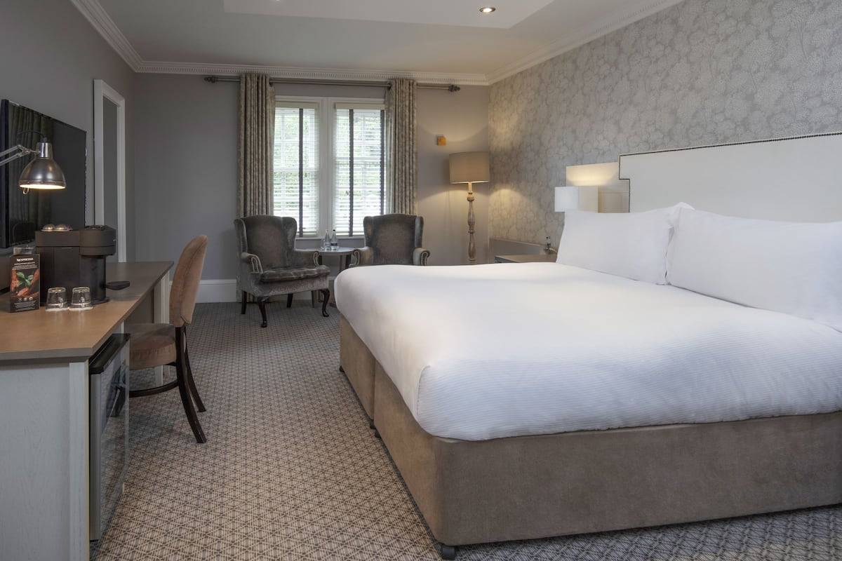 A hotel room with a large bed, a desk with a lamp, a chair, a small table, and a window with curtains.