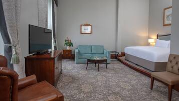 A hotel room with a large bed, a sofa, a television, a small table, and a decorative plant.