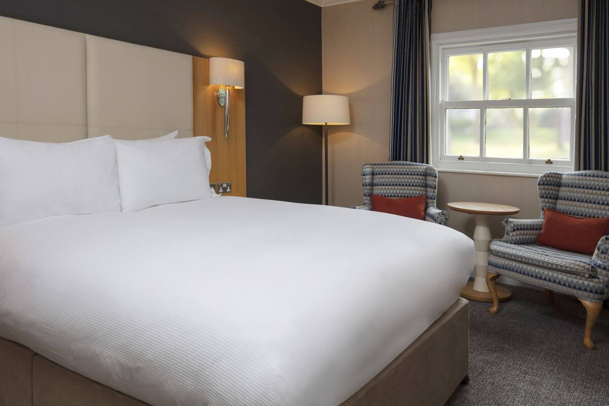 A hotel room with a large bed, a bedside lamp, a small table, two patterned chairs, and a window with curtains.