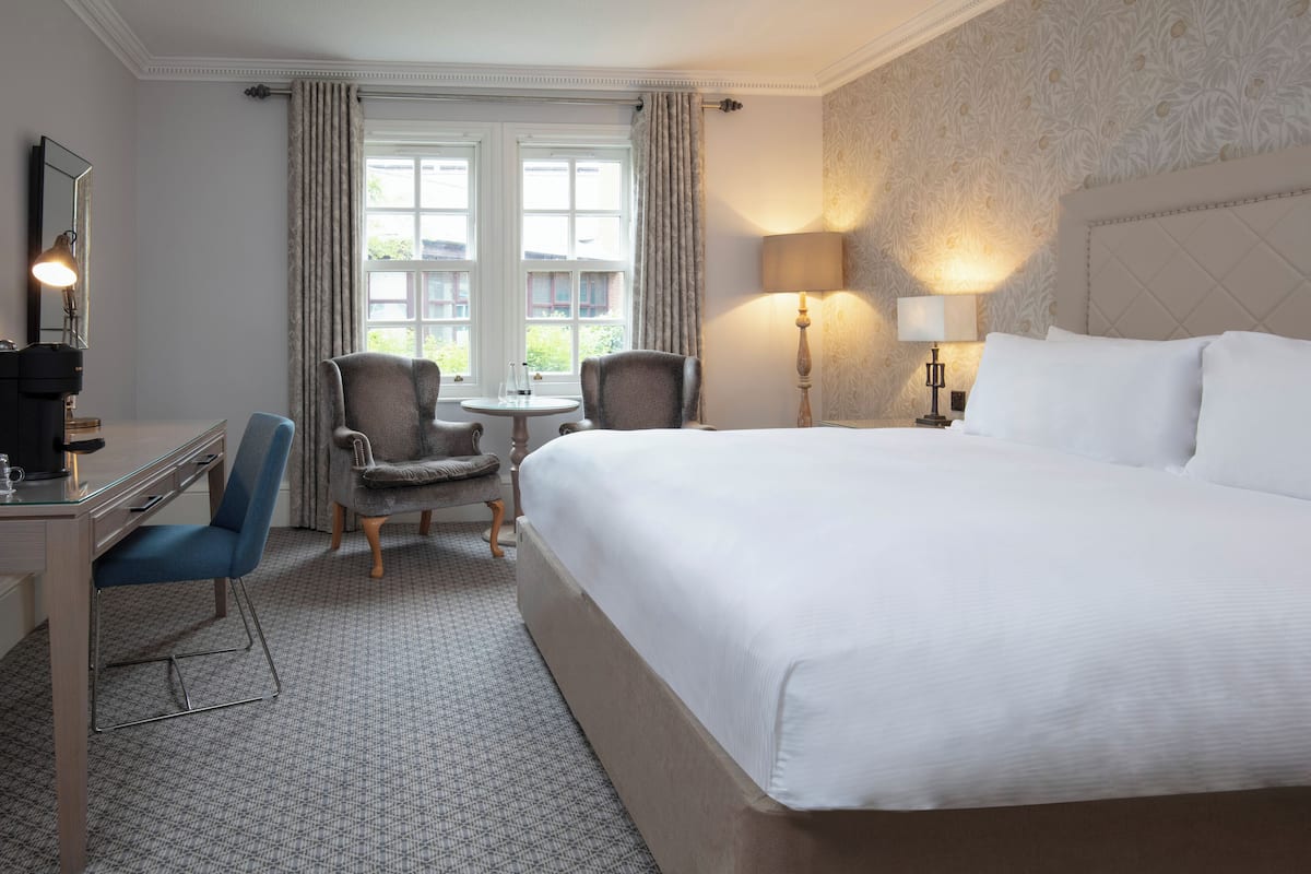 A hotel room with a large bed, a desk, two chairs, a small table, and a window with curtains.