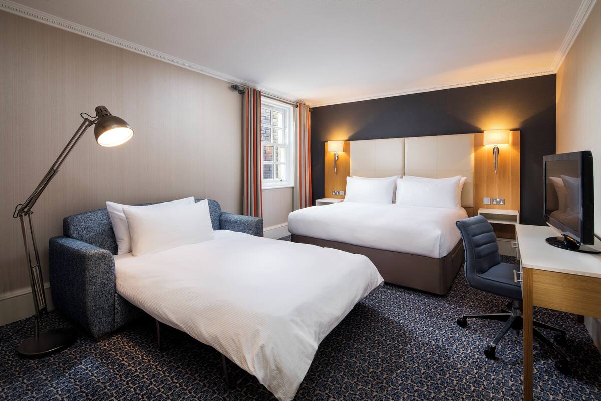 A hotel room with two beds, a desk, a chair, a lamp, and a television.