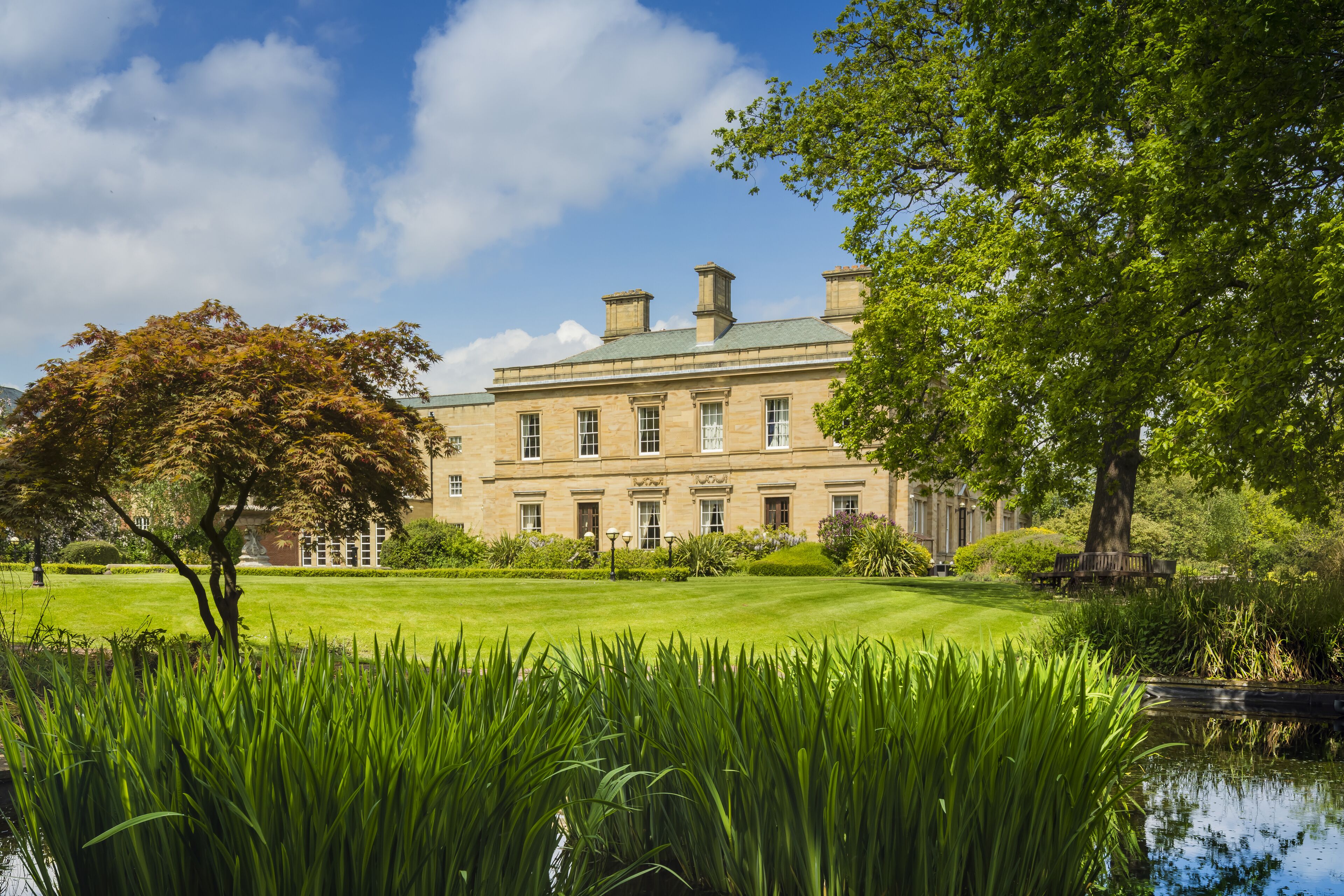 A large, historic building with a well-maintained lawn and a pond in the foreground.
