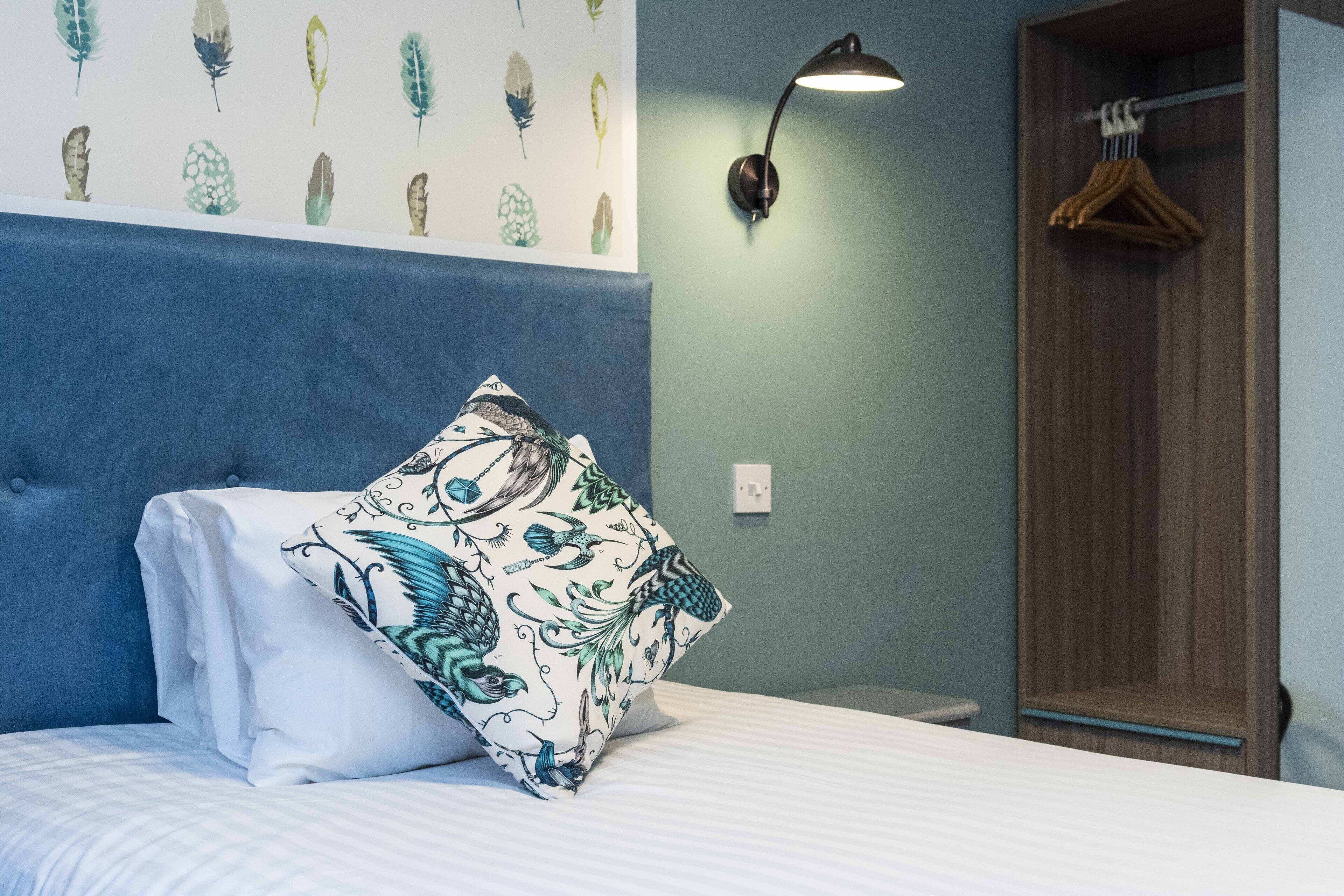 A neatly made bed with a decorative pillow, a wall-mounted light fixture, and a wooden wardrobe in a room with a blue accent wall.