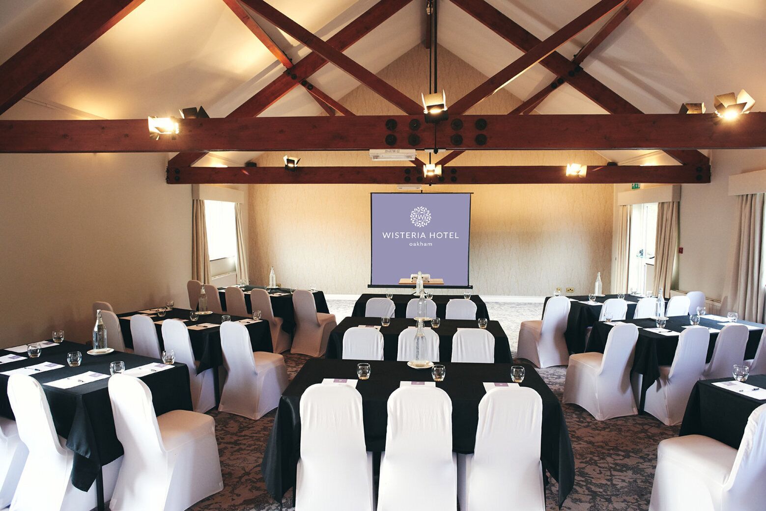 Une salle de conférence à l’hôtel Wisteria, avec des tables disposées pour une réunion, des chaises blanches et un écran de projection en arrière-plan.