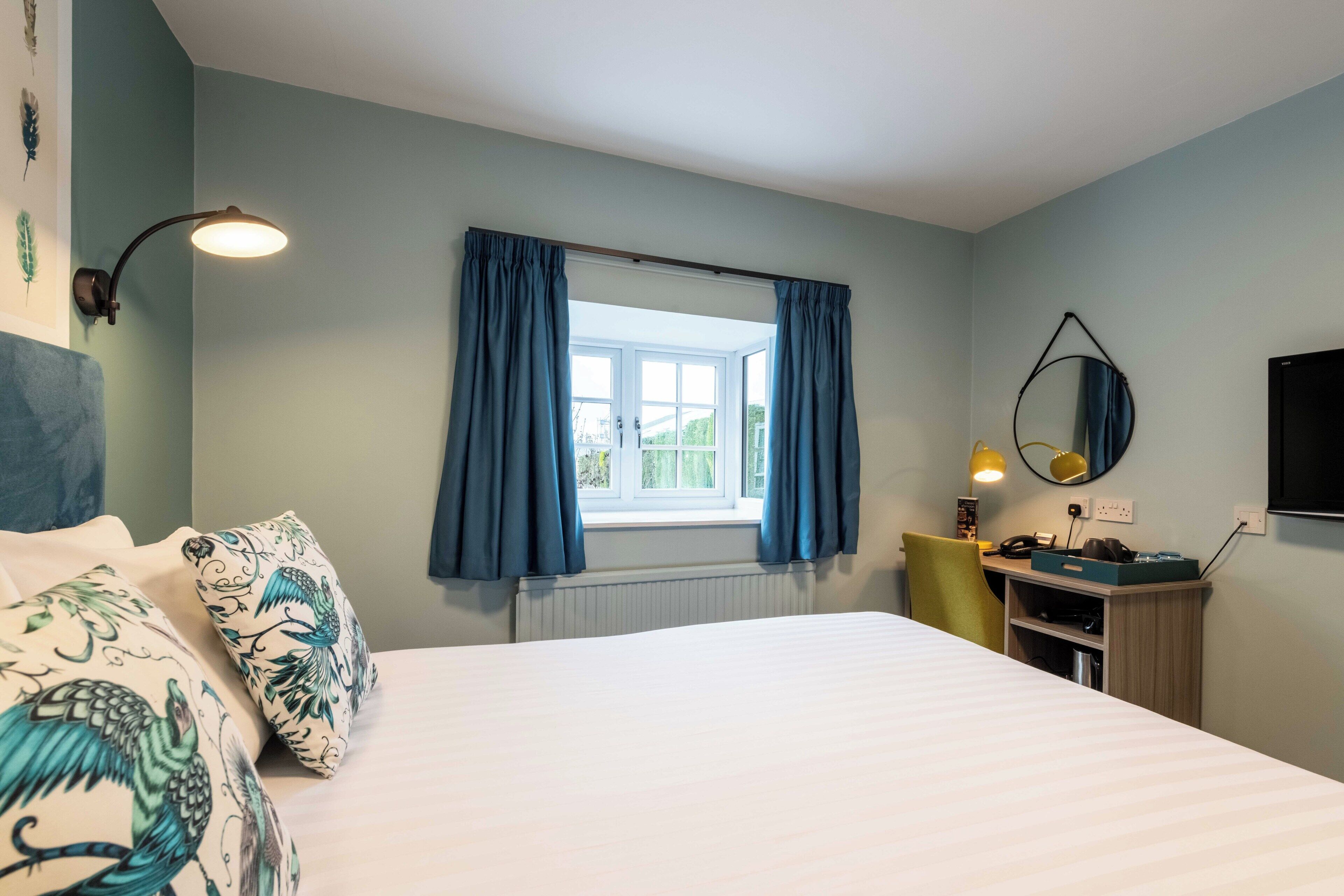 A bedroom with a bed, a desk, a chair, a window with curtains, a wall-mounted lamp, and a mirror.