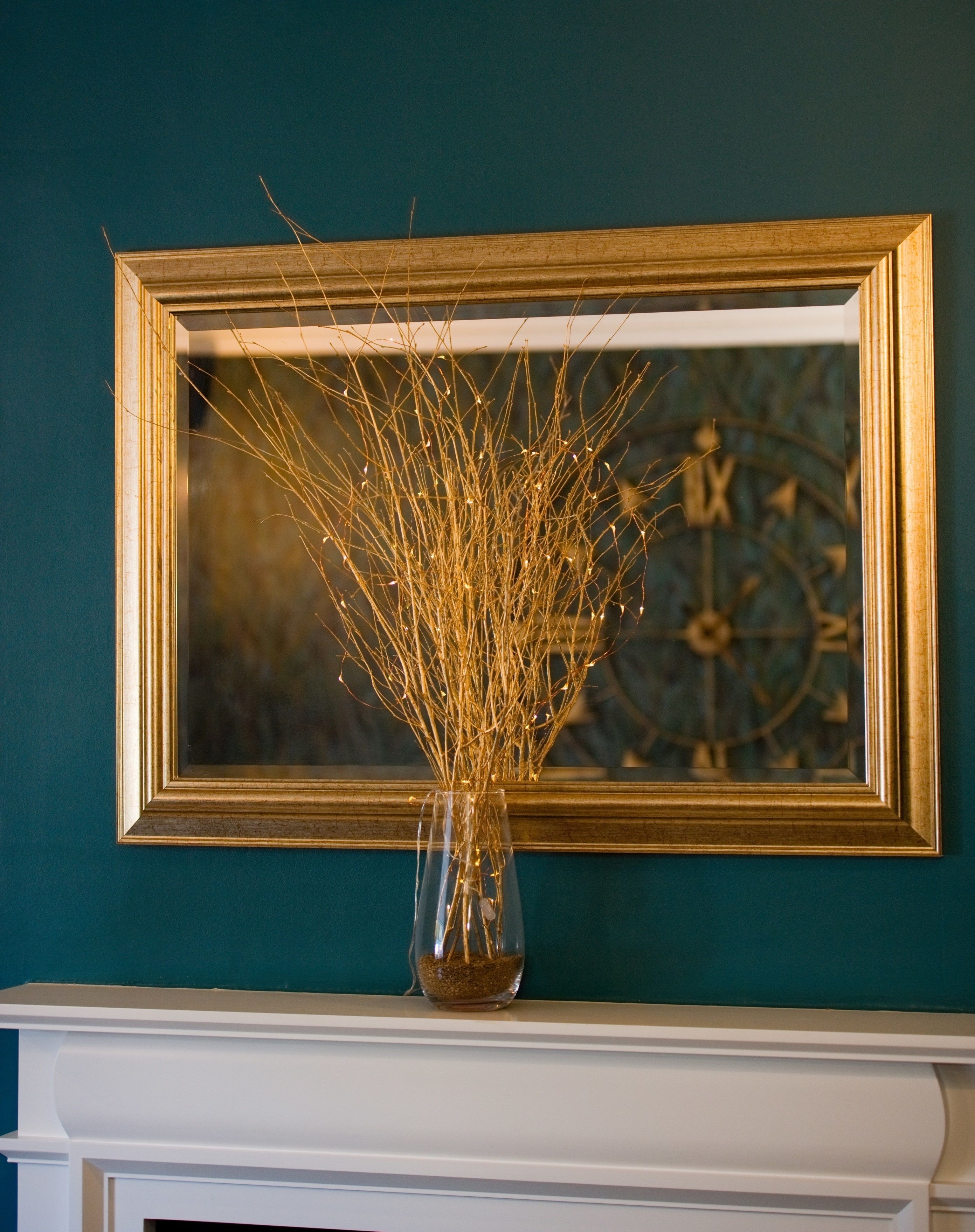 Eine Vase mit goldenen Zweigen auf einem Kaminvorsatz vor einer türkisfarbenen Wand, darüber ein gerahmtes Kunstwerk.