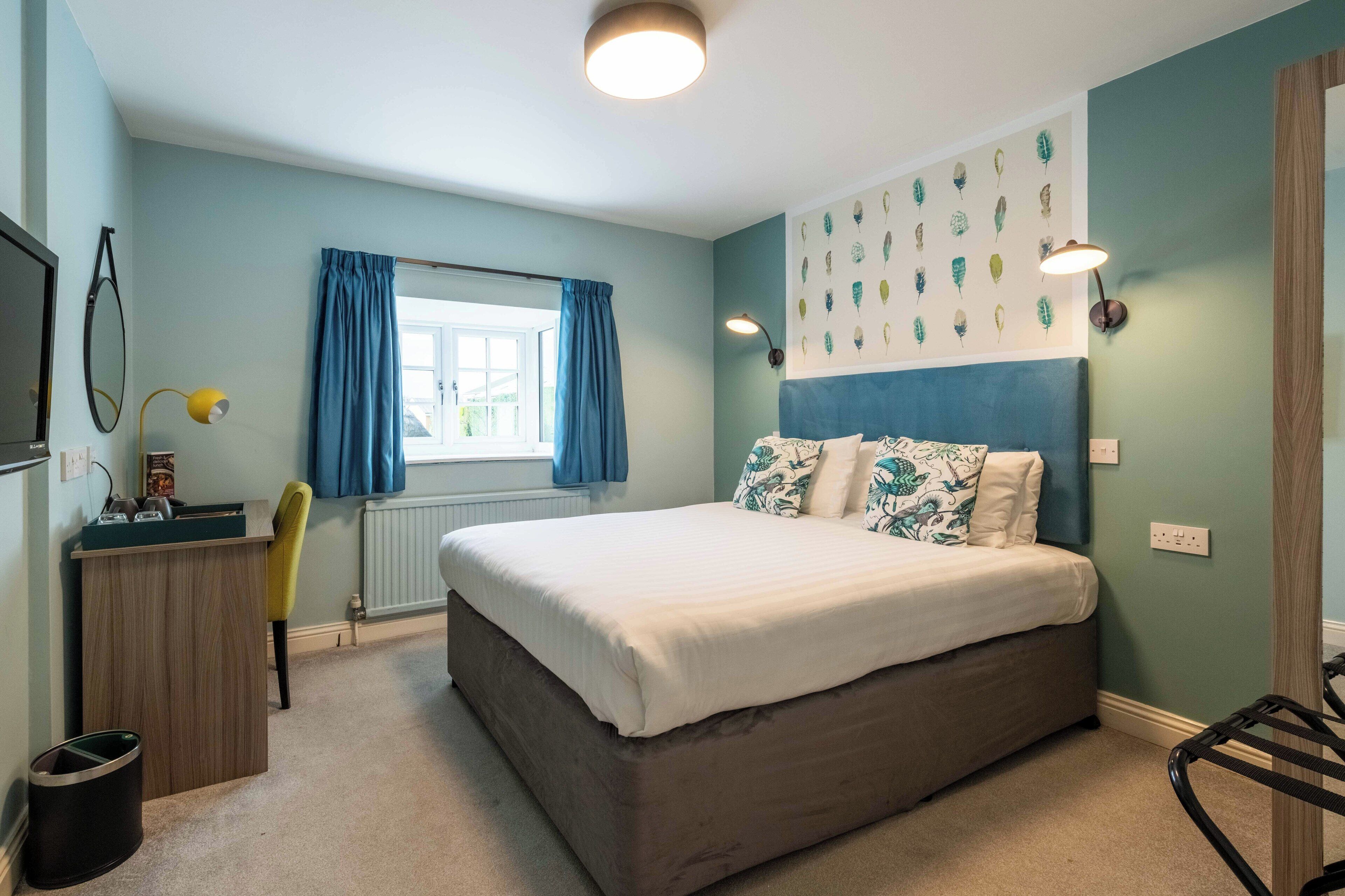 A bedroom with a bed, a desk, a chair, a radiator, a window with curtains, and wall art.
