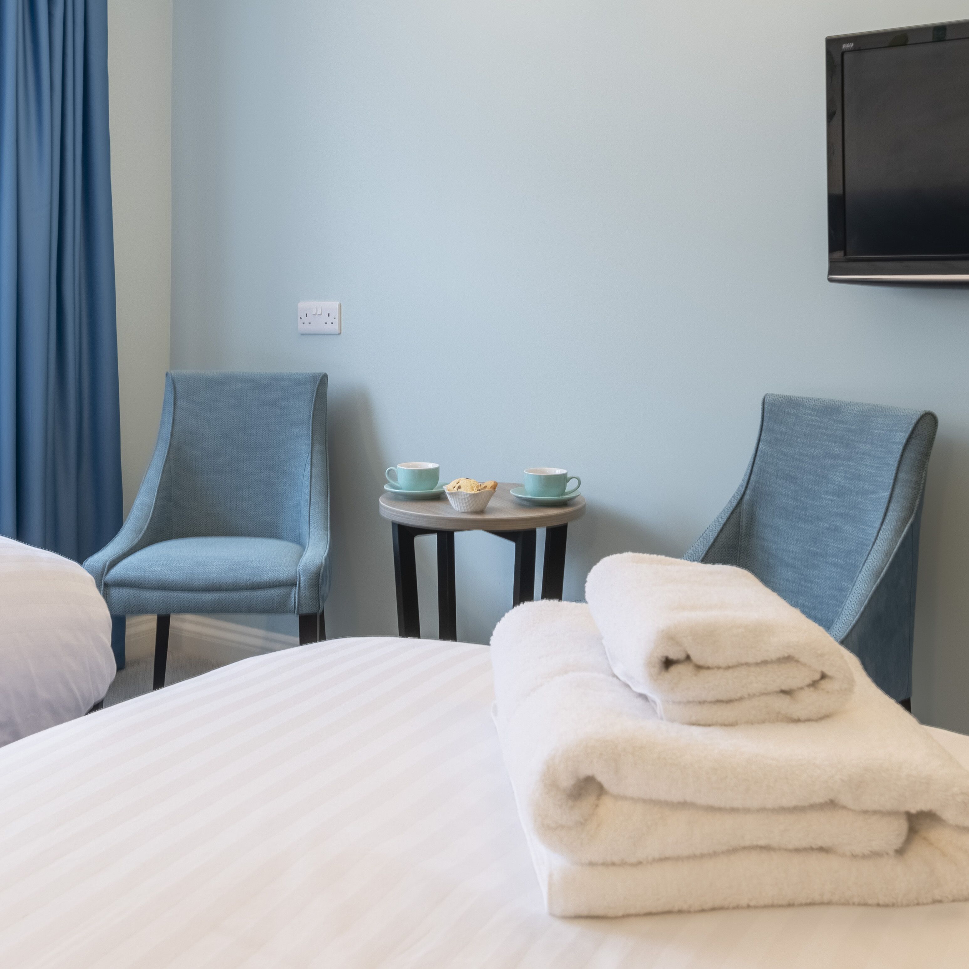A hotel room with a bed, two armchairs, a small table with cups and a bowl, and a TV mounted on the wall.