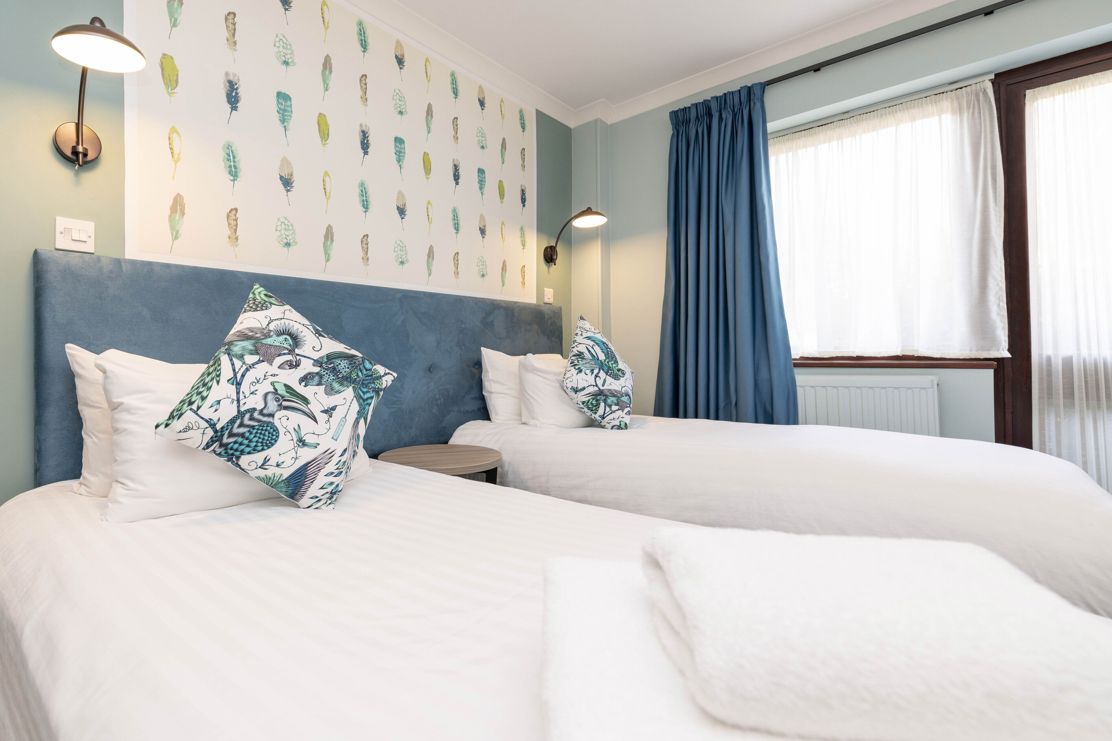 A hotel room with two beds, a headboard, wall art, a lamp, and a window with curtains.