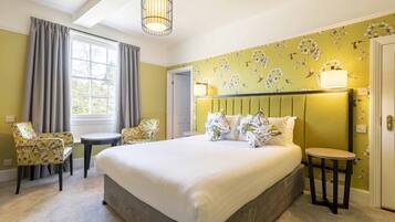 Un dormitorio con una cama grande, dos sillones, una mesita, una ventana con cortinas y un papel tapiz con motivos florales.
