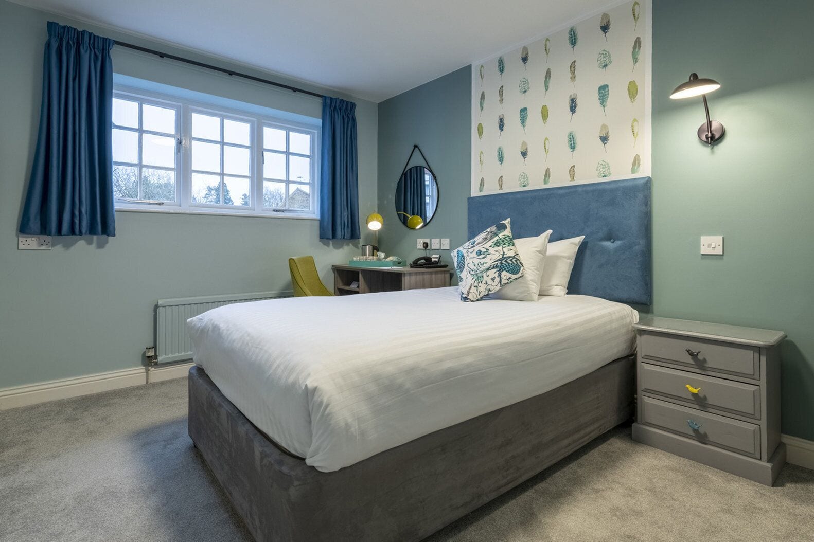 Een slaapkamer met een groot bed, een bureau, een stoel, een bedlampje en een muur met bladmotieven.