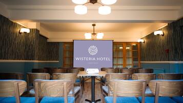 A conference room with a projector screen displaying "Wisteria Hotel Oakham" logo, wooden chairs with blue cushions, and a ceiling fan.