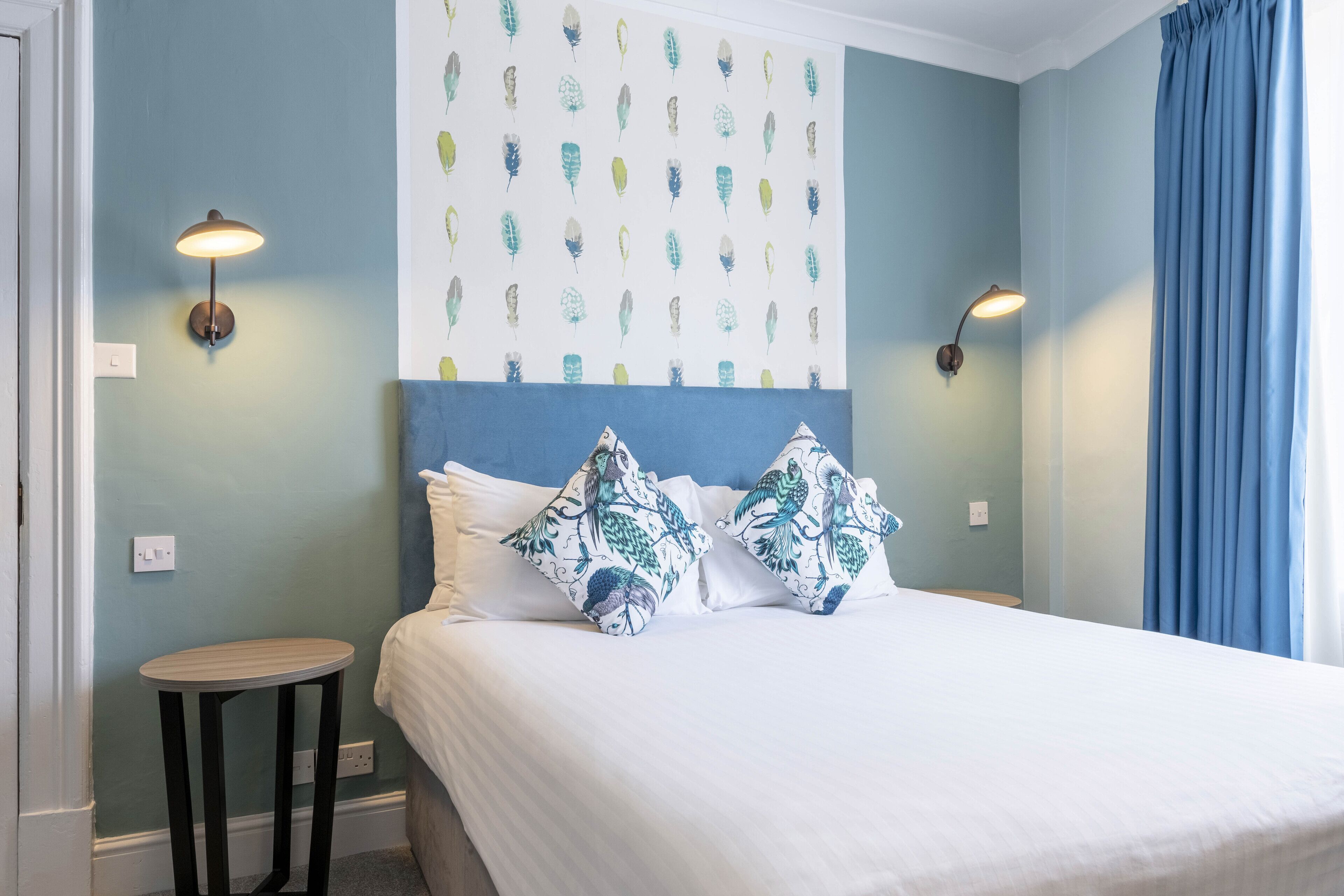 A bedroom with a bed, two wall-mounted lamps, a small round table, and a patterned wall.