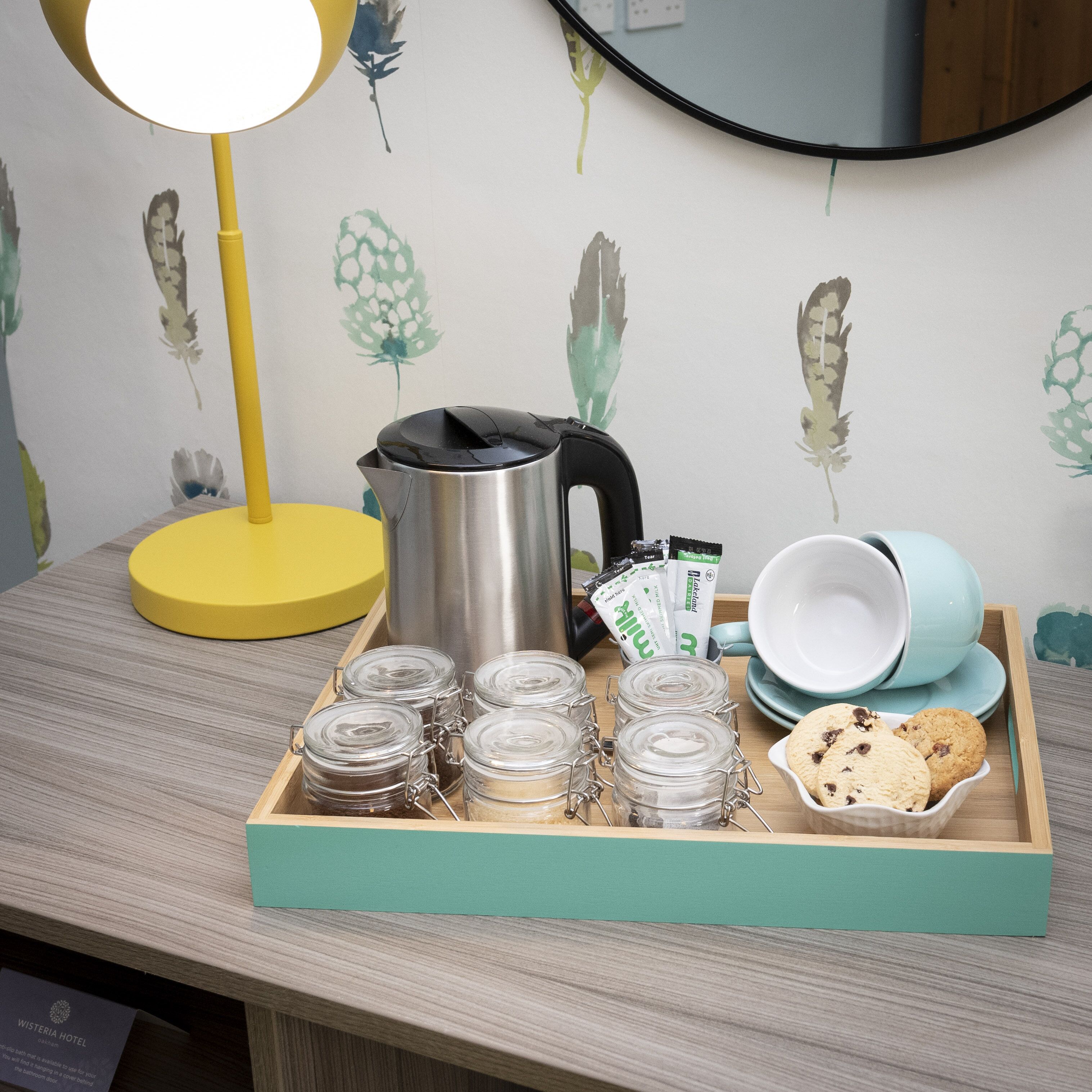 A wooden tray with jars, a coffee pot, tea packets, and cookies on a table.