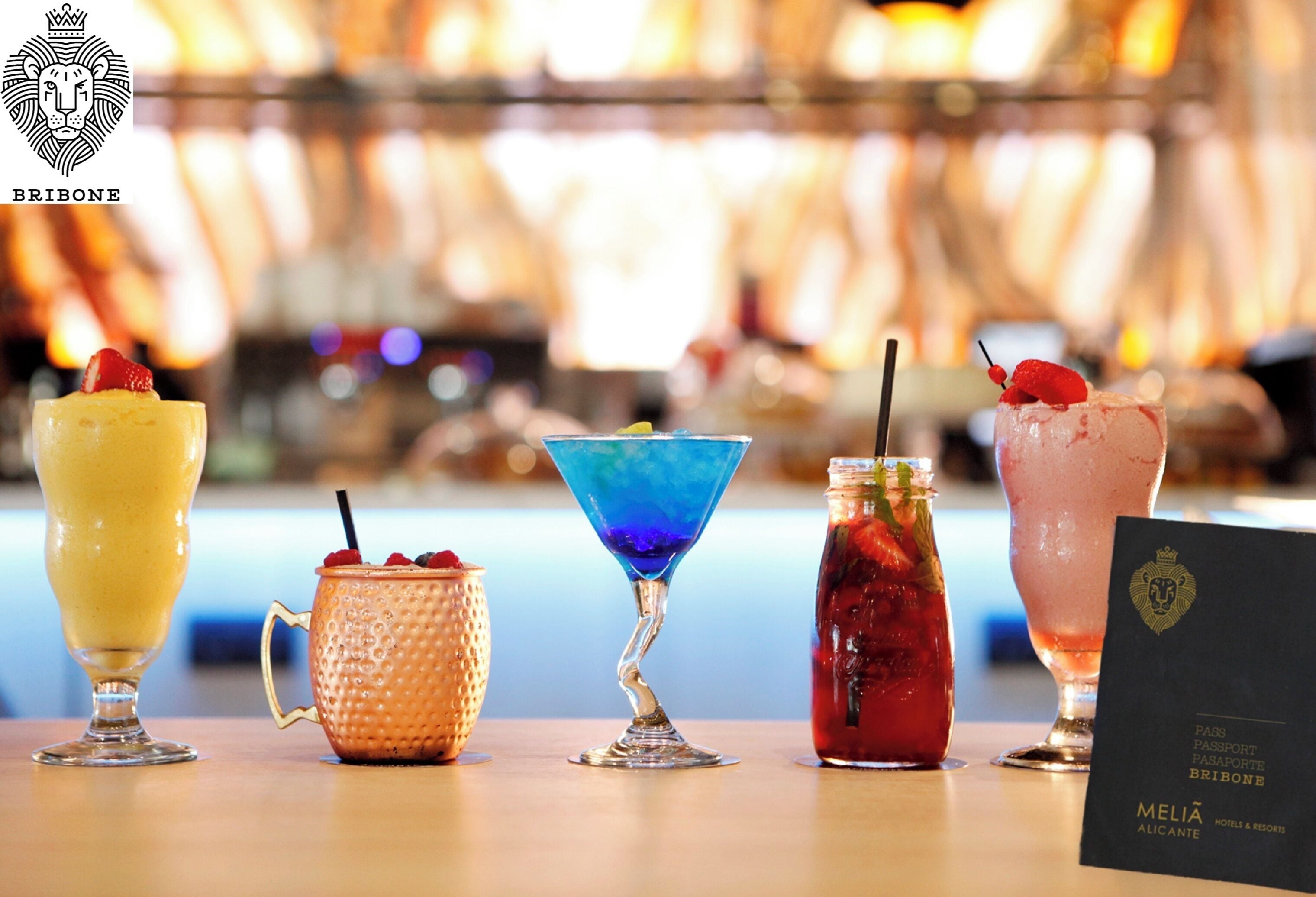 Variedad de bebidas coloridas sobre una barra, incluyendo una bebida amarilla, una azul, una roja y una rosa, cada una adornada con fruta.