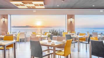 Un restaurant avec des tables et des chaises disposées pour accueillir les clients, de grandes fenêtres offrant une vue sur le coucher de soleil sur l’eau.