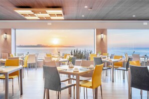 A restaurant with tables and chairs arranged for dining, large windows offering a view of the sunset over the water. - Melia Alicante (Alicante)