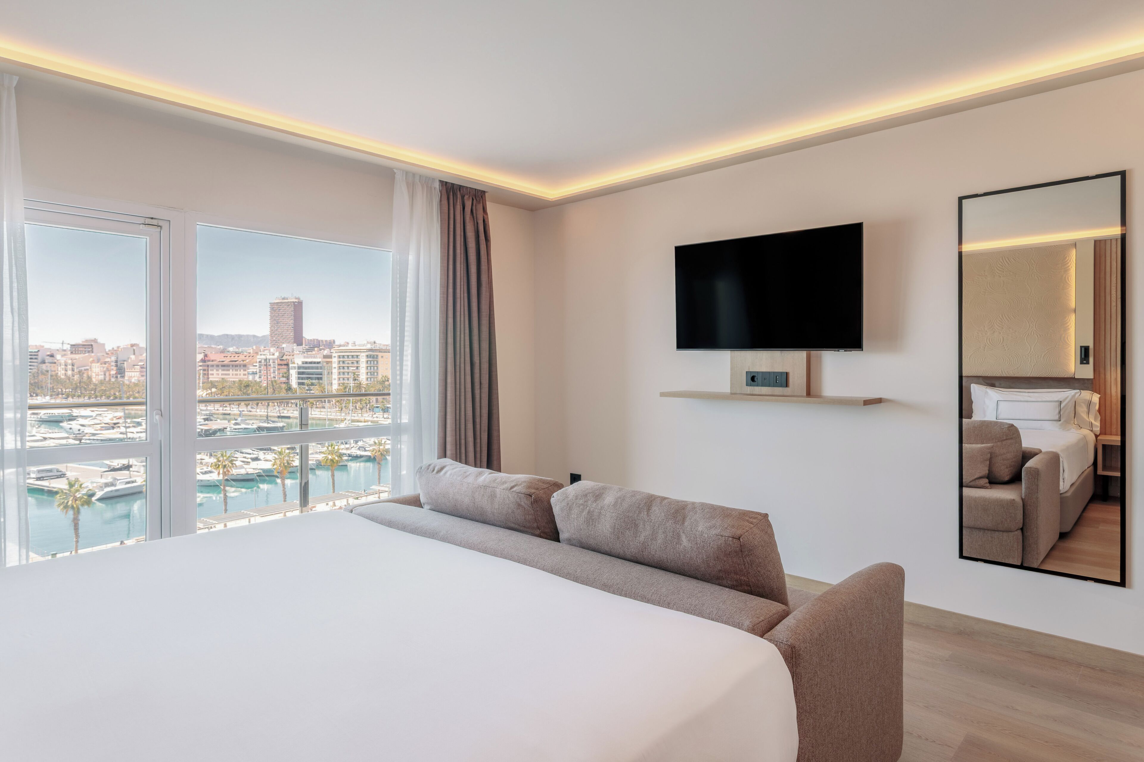 A hotel room with a large bed, a TV mounted on the wall, a view of a marina, and a cityscape through the window.