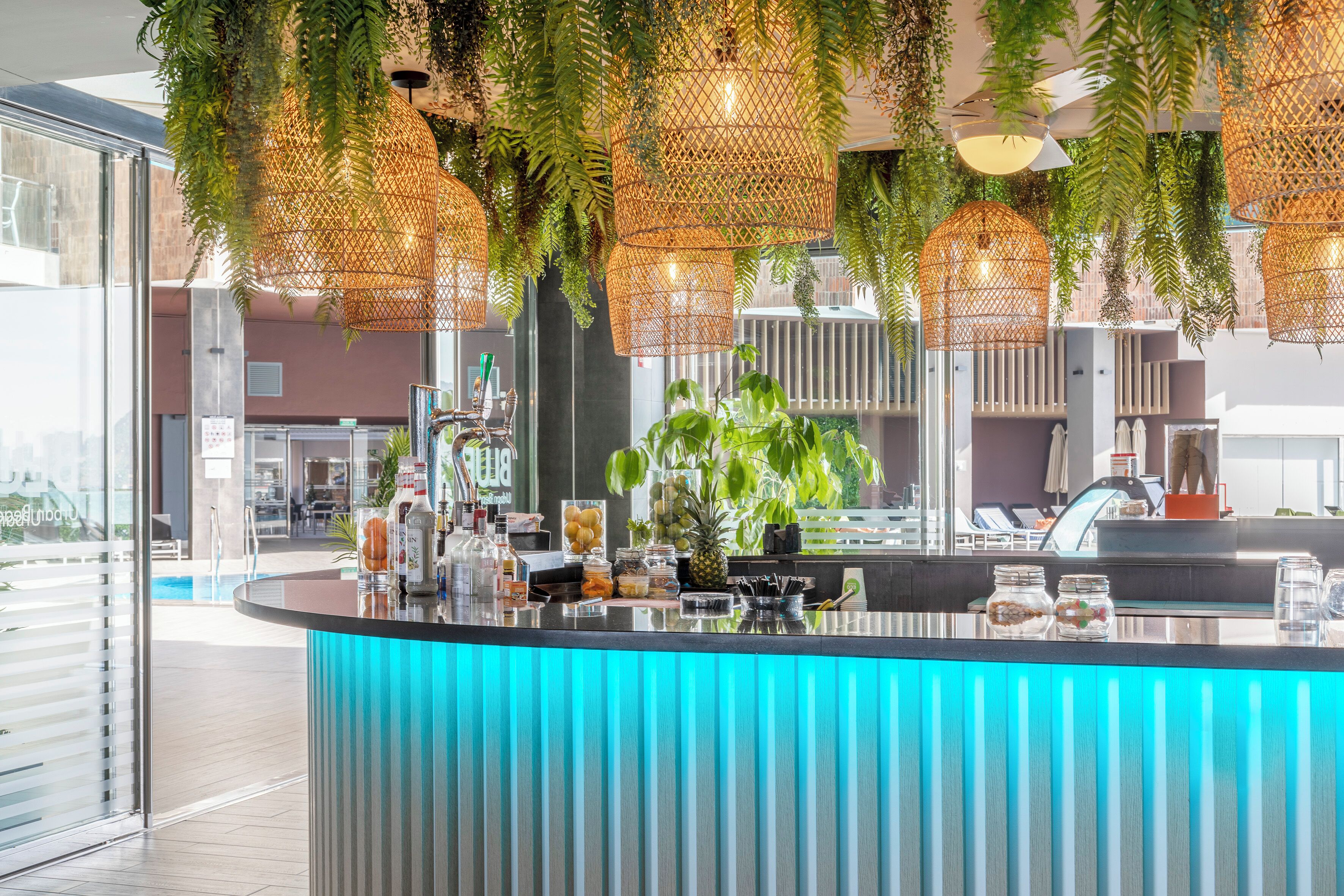 A modern bar area with hanging wicker lamps, a counter with various items, and a view of the pool outside.