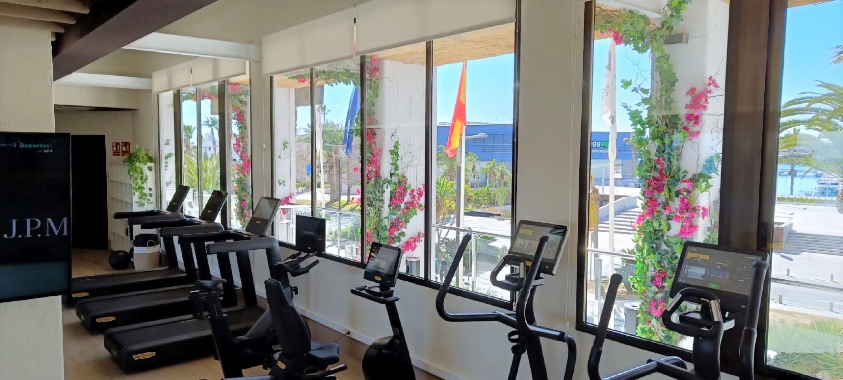 A gym with treadmills and exercise bikes, large windows with plants, and a J.P.M sign.