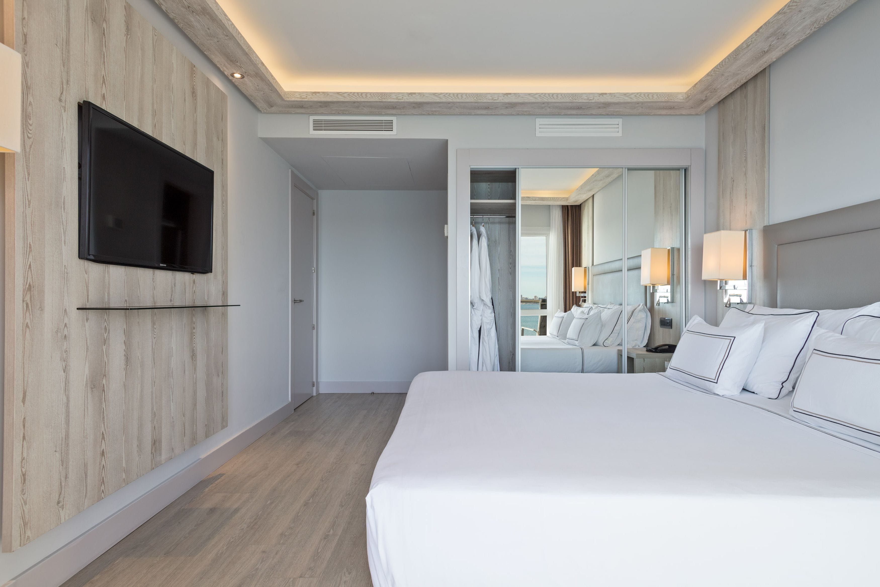 Una habitación de hotel moderna con una cama grande, un televisor de pantalla plana montado en una pared de madera, un vestidor y un baño visible a través de una puerta abierta.