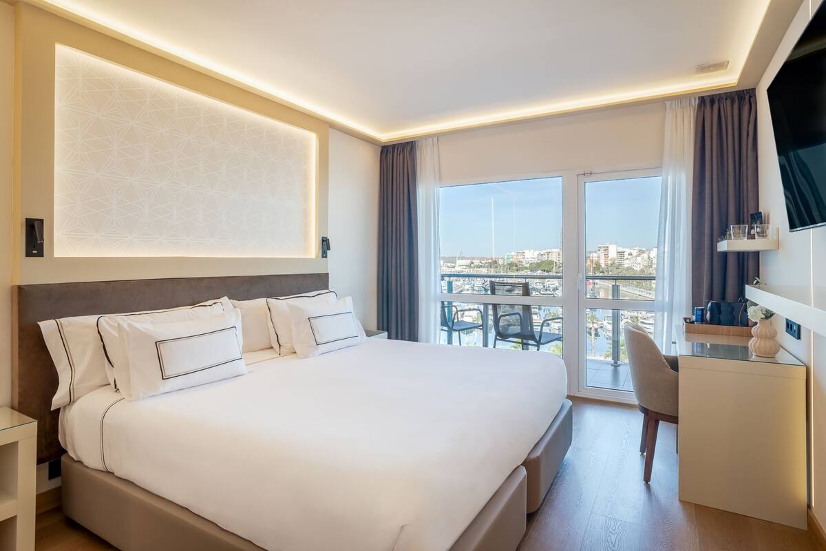 A modern hotel room with a large bed, a desk, and a balcony with a city view.