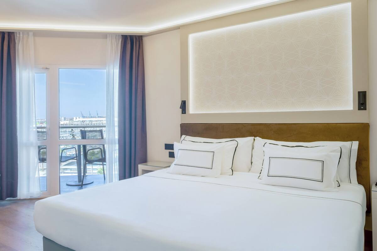 A hotel room with a large bed, a nightstand, a wall-mounted light, and a view of a cityscape through a glass door.
