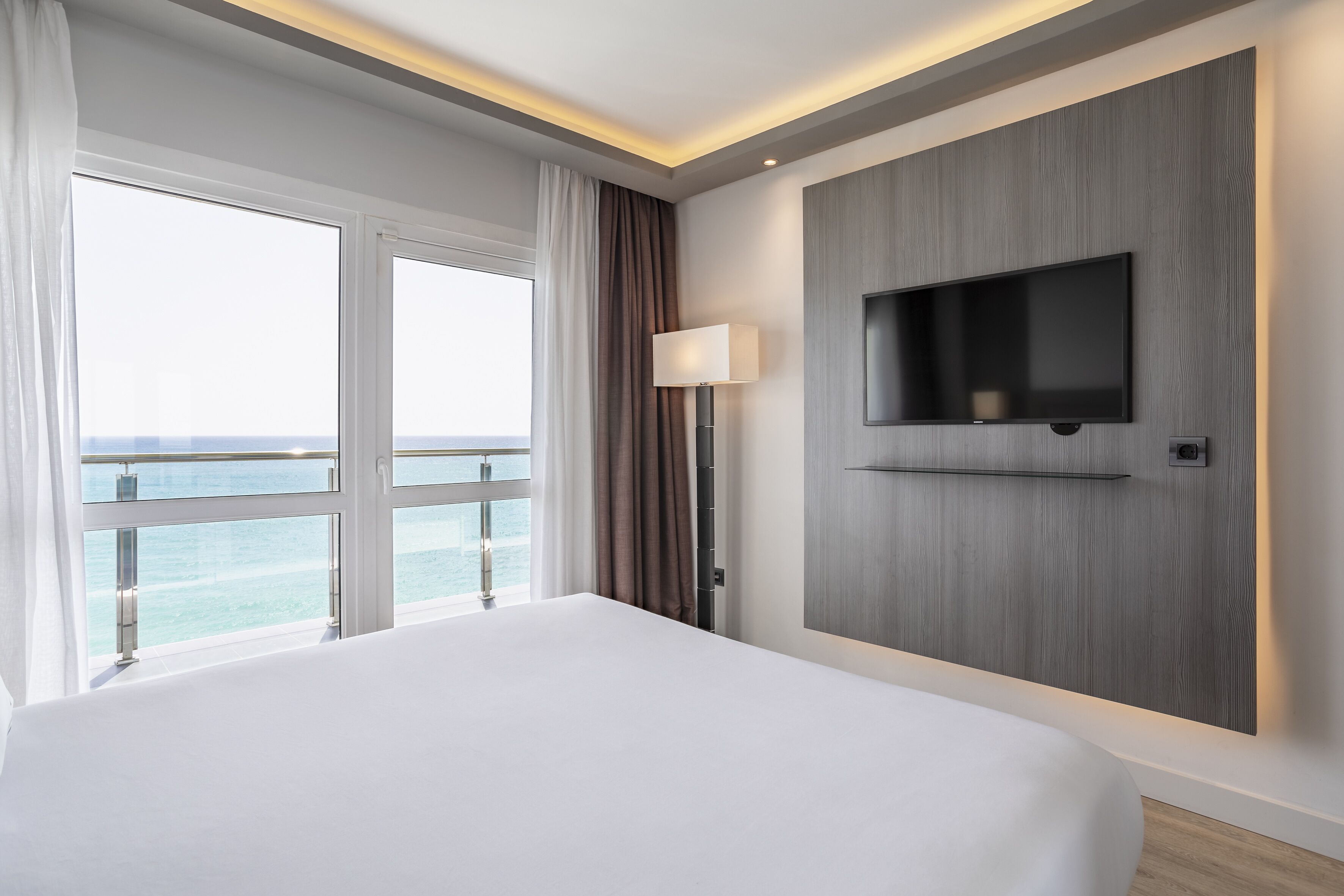 Een moderne hotelkamer met een groot bed, een aan de muur bevestigde televisie en uitzicht op de oceaan door grote ramen.