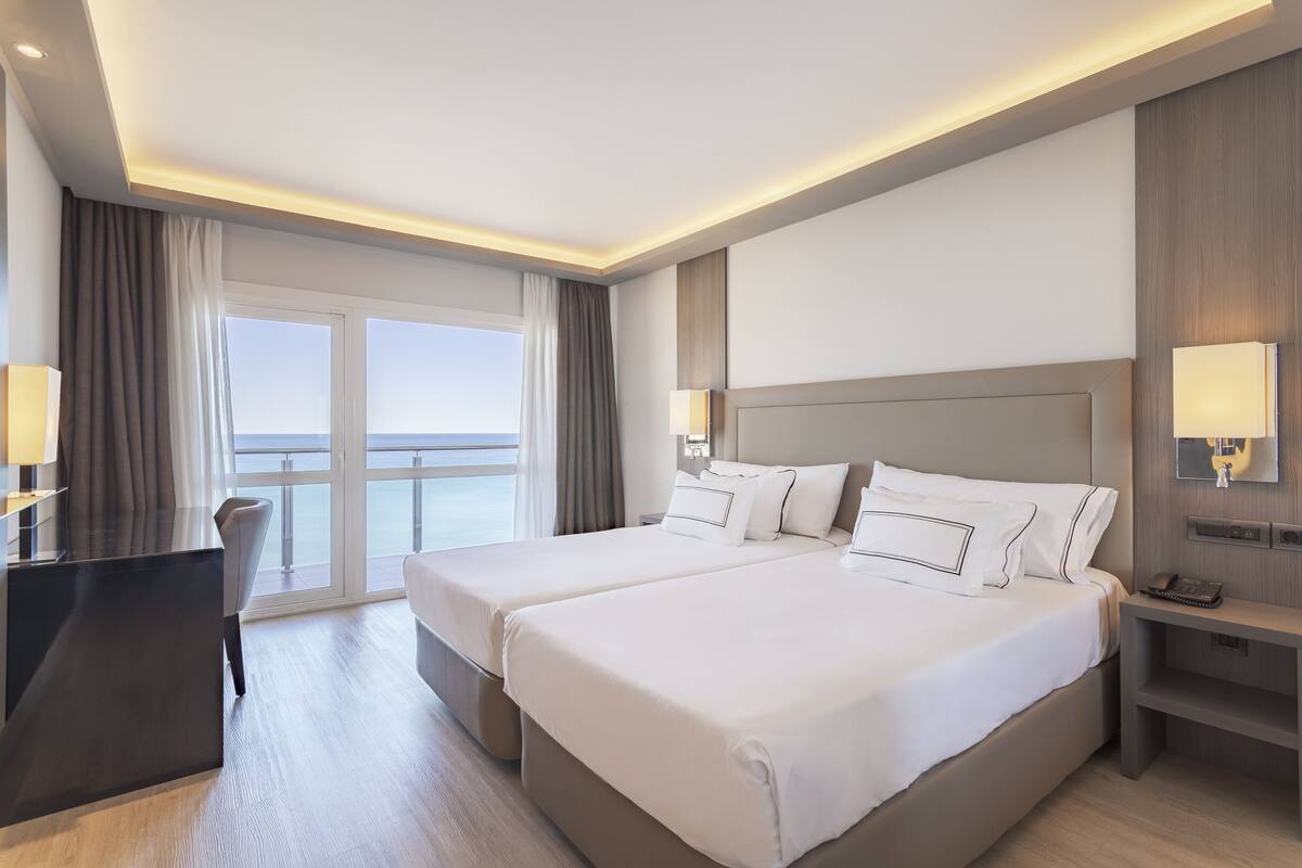 A hotel room with two beds, a desk, and a view of the sea.