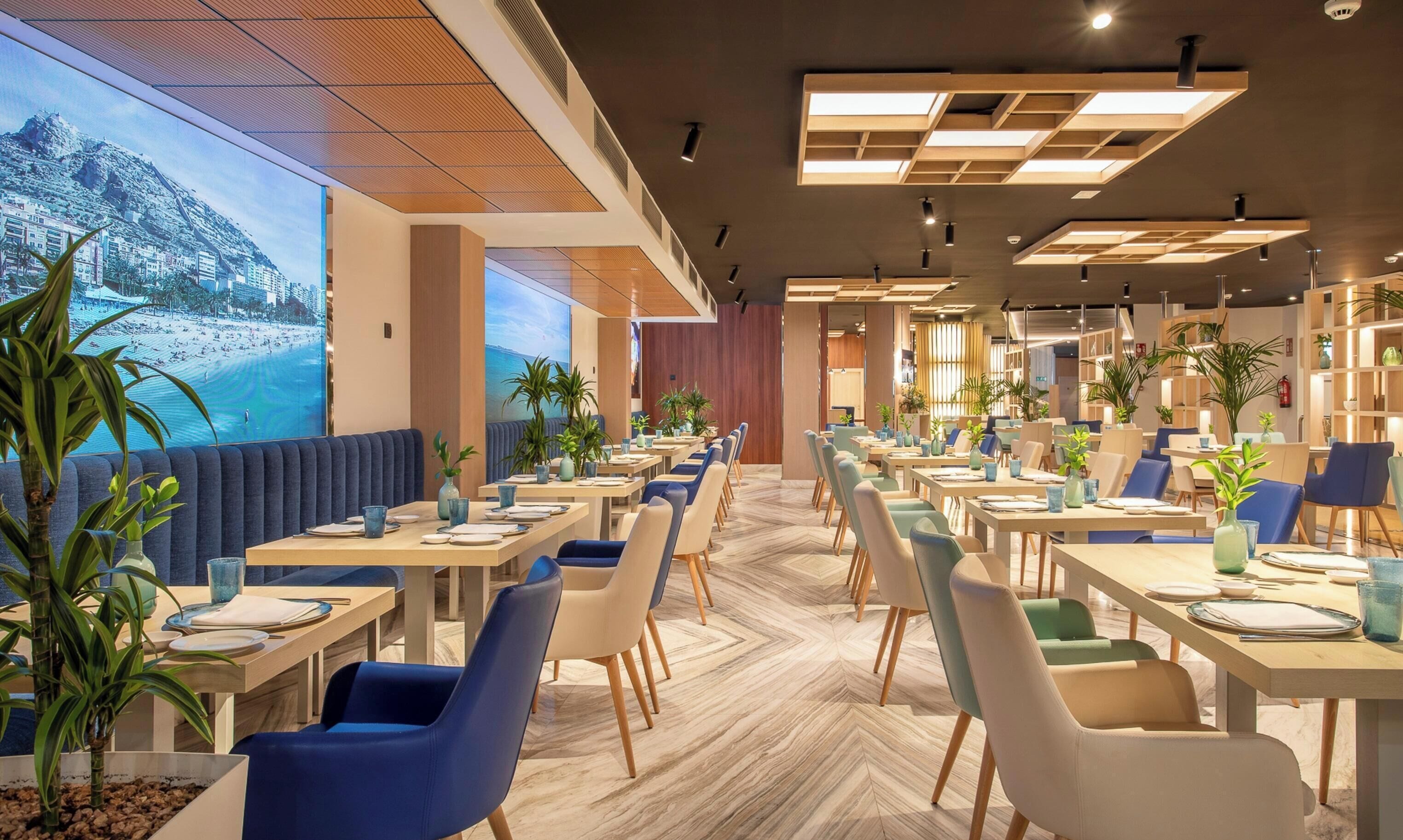Een modern restaurant met uitzicht op een strand en bergen, met houten tafels, blauw-witte stoelen en potplanten.