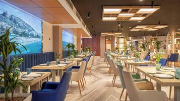 Een modern restaurant met uitzicht op een strand en bergen, met houten tafels, blauw-witte stoelen en potplanten.