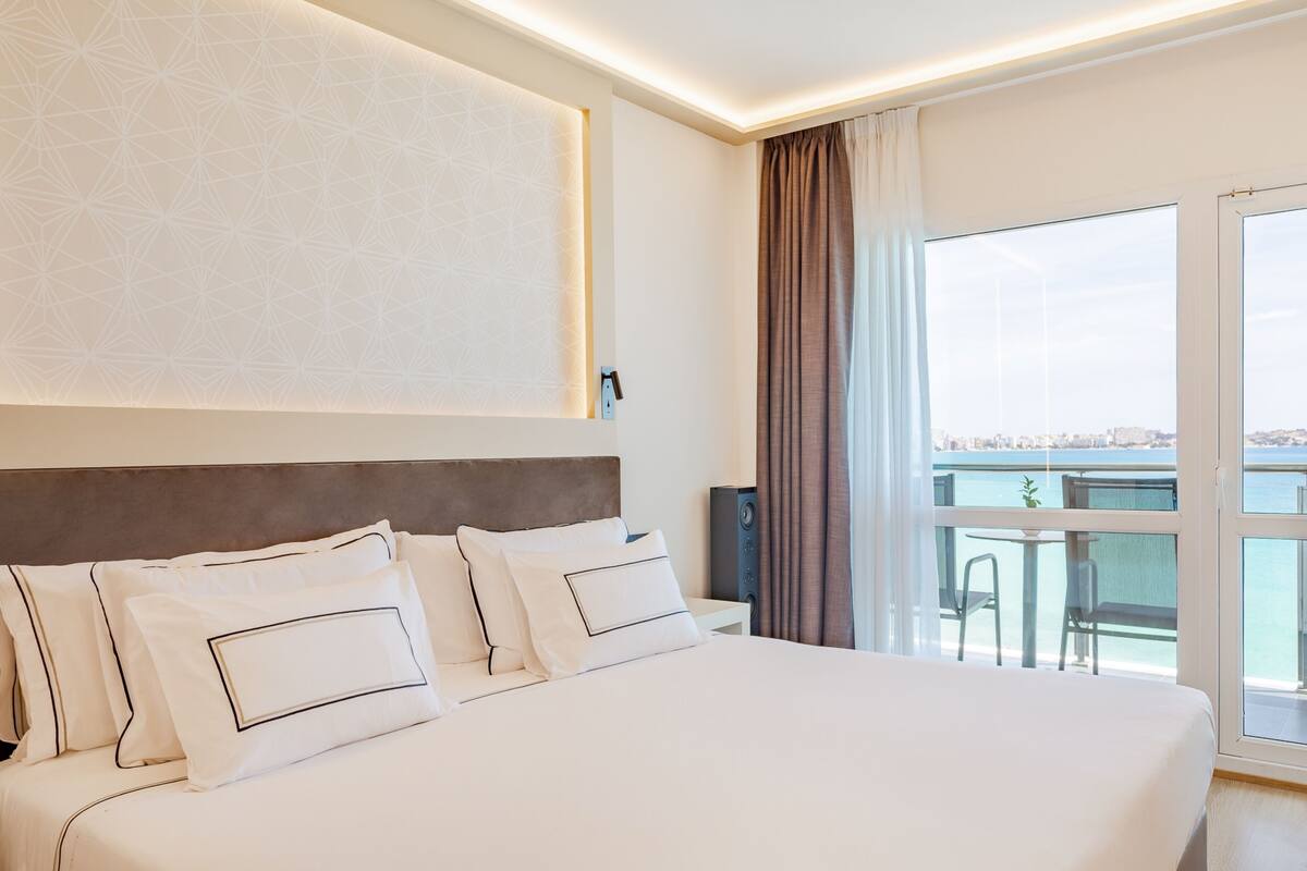 A hotel room with a large bed, a view of the city through the window, and a balcony with a table and chairs.