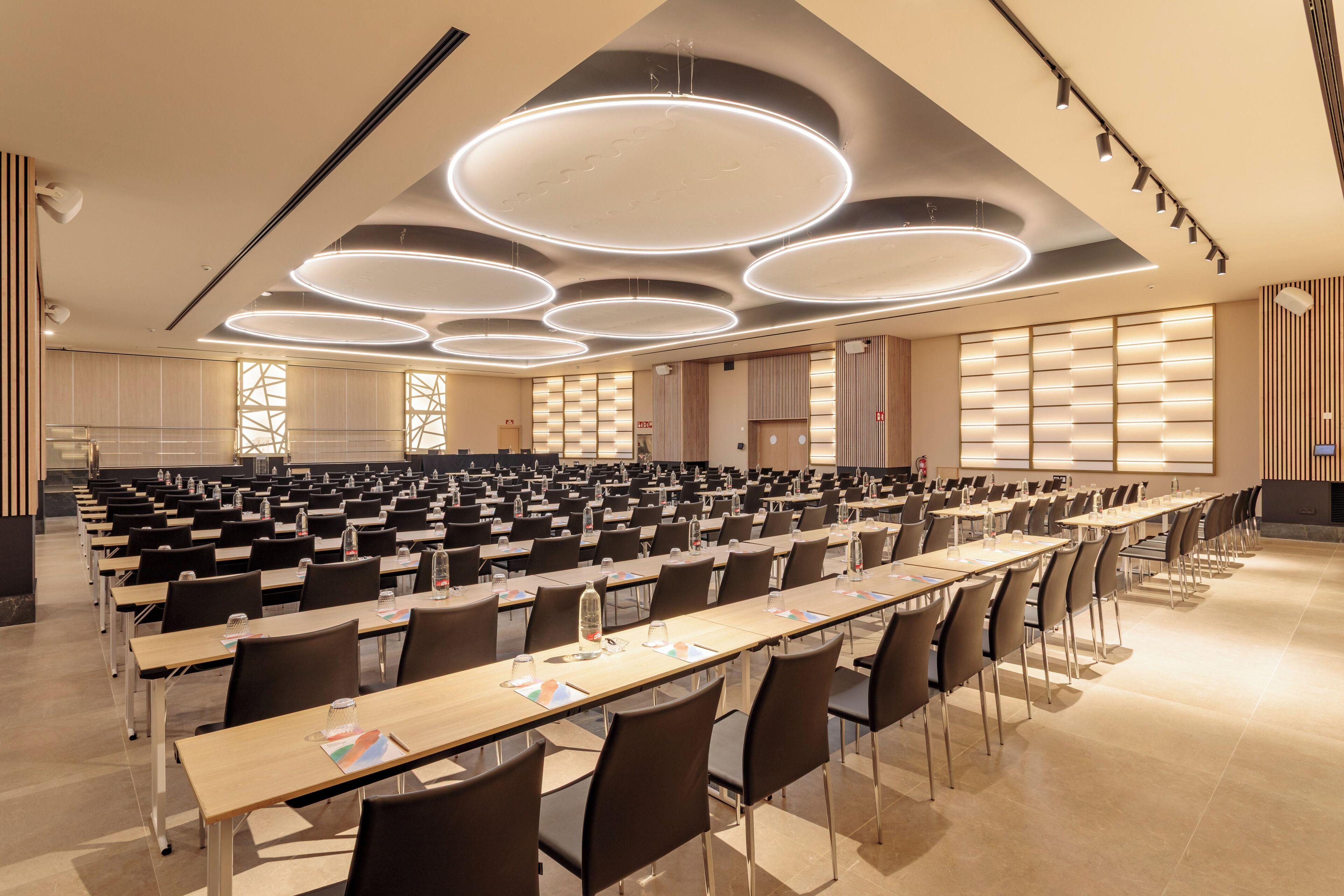 Een grote congreszaal met meerdere rijen tafels en stoelen, voorzien van waterflesjes en notitieblokken.