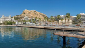 A marina with yachts docked, a large building with a hill in the background, and a hotel with palm trees. - Melia Alicante (Alicante)