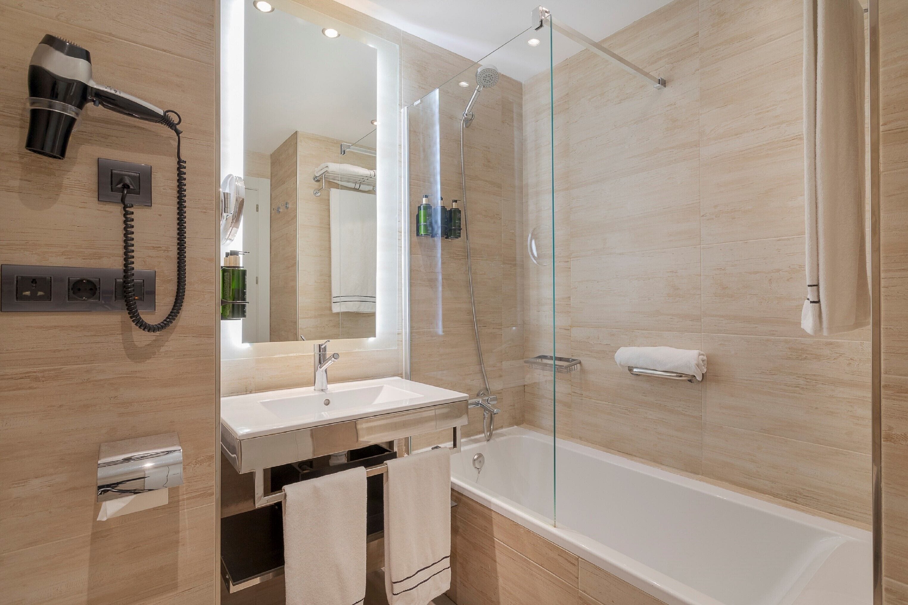Un baño moderno con ducha acristalada, secador de pelo montado en la pared y lavamanos blanco.
