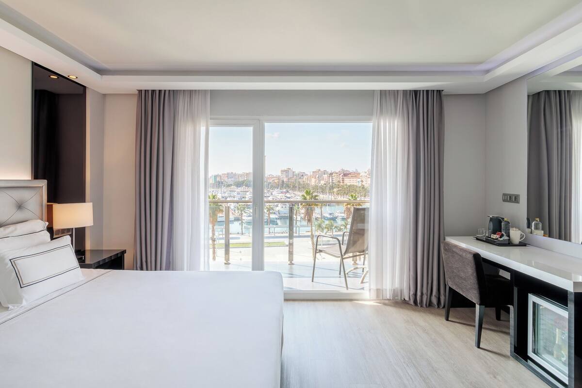 A hotel room with a bed, a desk, a chair, and a large window offering a view of a marina and cityscape.