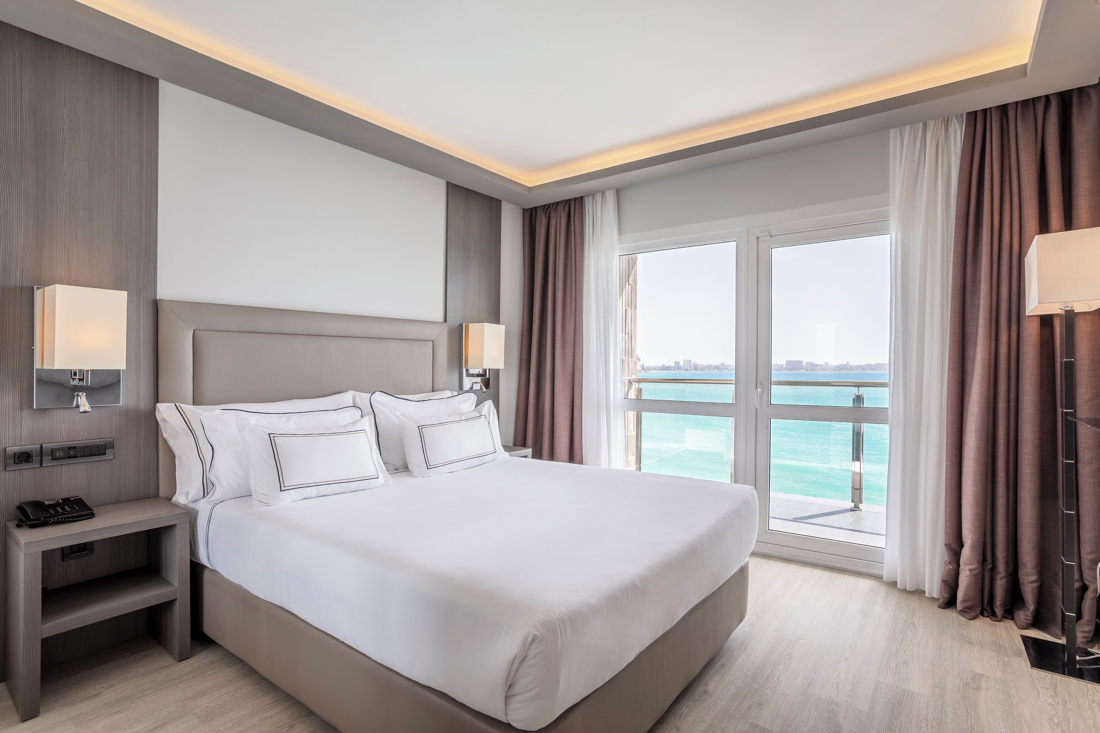 Ein modernes Hotelzimmer mit einem großen Bett, Nachttischlampen, einem Beistelltisch und Meerblick durch ein großes Fenster.
