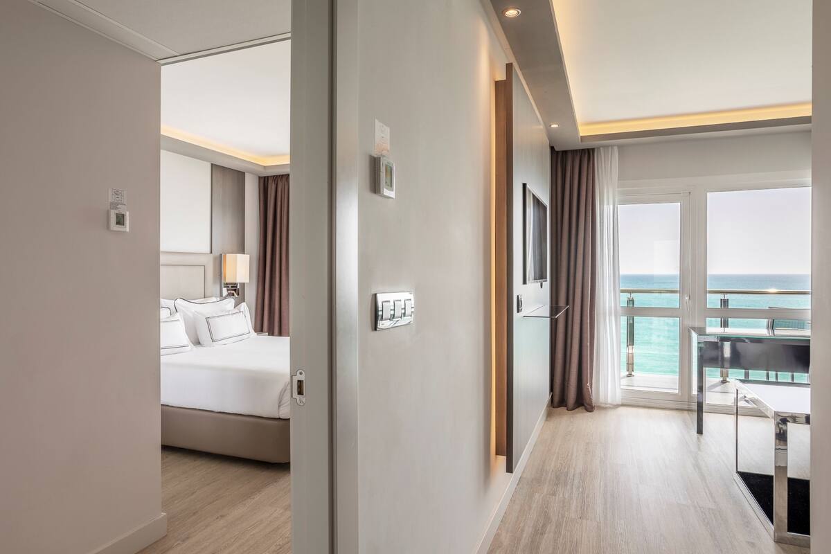 A hotel room with a bed, bedside table, lamp, and a view of the sea through large windows.