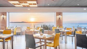 A dining area with tables and chairs, large windows with a view of the sunset over the water. - Melia Alicante (Alicante)