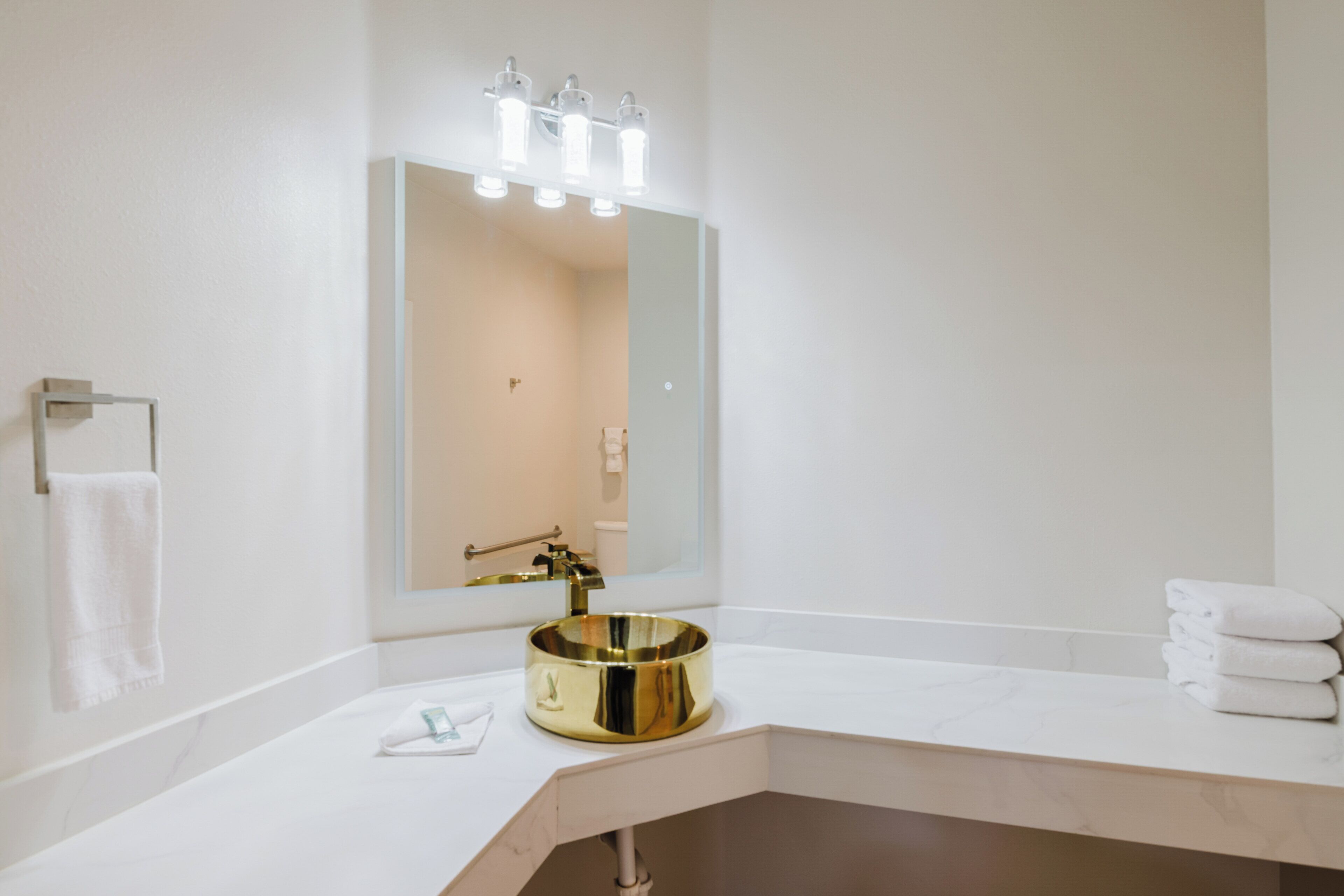 Un baño con tocador blanco, lavamanos dorado, un espejo grande con iluminación y un perchero para toallas.