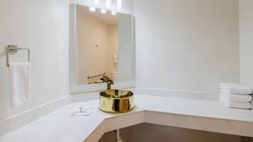 Un baño con tocador blanco, lavamanos dorado, un espejo grande con iluminación y un perchero para toallas.