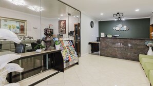 Un área de recepción con un separador de vidrio, mostrador de recepción, pared verde con un adorno circular, un reloj y una zona de estar con un sofá verde.