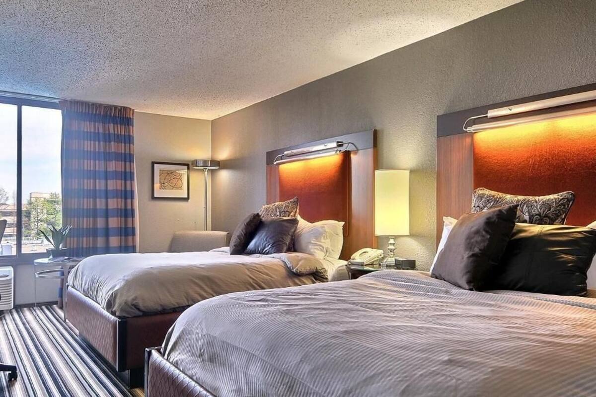 A hotel room with two beds, a desk, a chair, a window with curtains, and a view of the city.