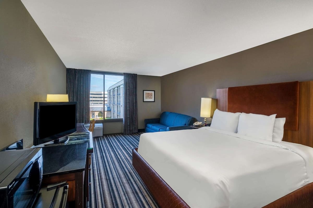 A hotel room with a large bed, a desk with a flat-screen TV, a blue sofa, and a window with a view of buildings.