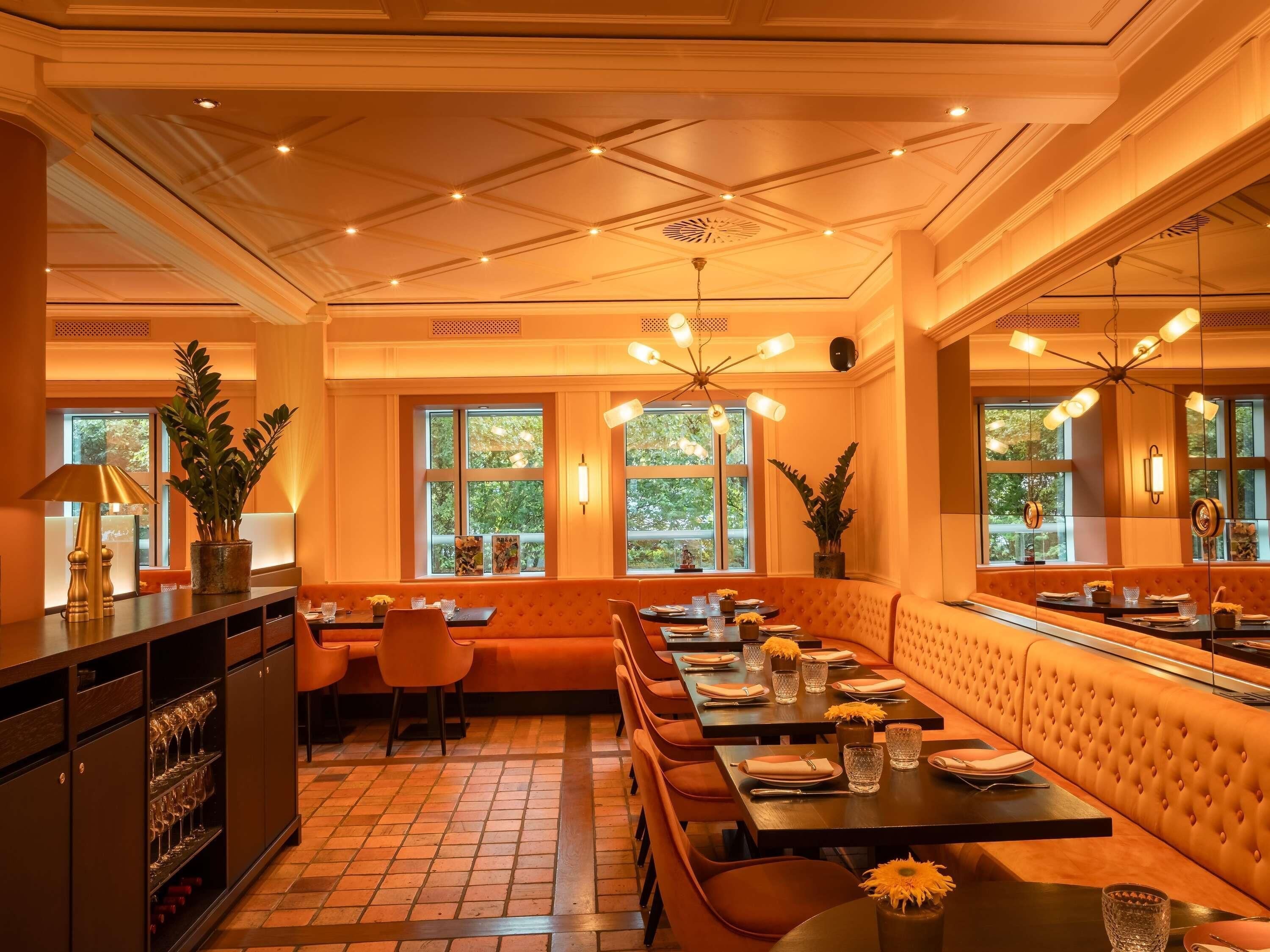 A restaurant with a warm interior, featuring a long dining table, cushioned seating, and large windows with natural light.