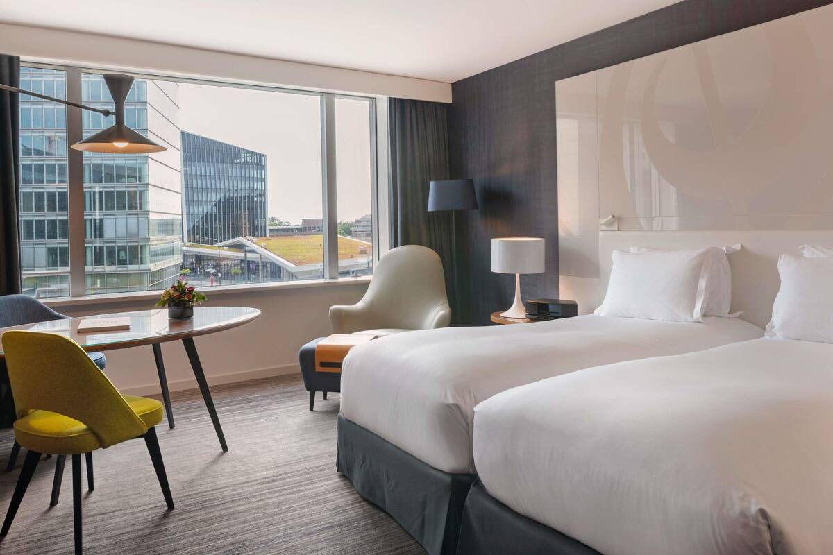 Une chambre d’hôtel avec un grand lit, un bureau avec une chaise, une lampe et une vue sur un immeuble par la fenêtre.